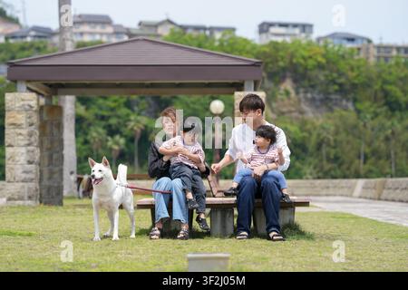 La famille profite d'un moment paisible dans le parc ensemble. Les enfants rient assis sur un banc avec leurs parents. Un chien blanc se tient à proximité, calme et attentif. Banque D'Images