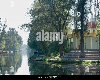 Canal pittoresque et tranquille avec des arbres verts luxuriants, de petites barques en bois et une belle chapelle traditionnelle blanche à dôme rouge se reflétant dans des eaux calmes et sereines Banque D'Images
