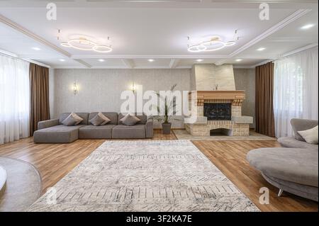 Salon lumineux avec un canapé d'angle gris, un tapis à motifs et une cheminée en pierre. La lumière naturelle filtre à travers les rideaux transparents. Deux lustres modernes Banque D'Images