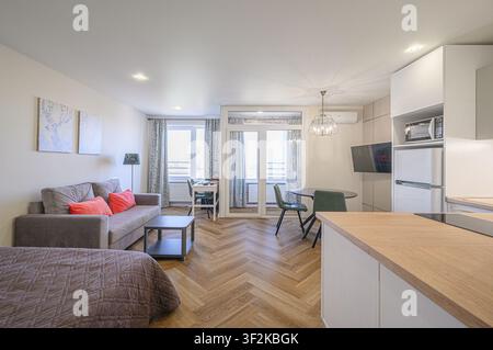 appartement studio lumineux avec des parquets en bois à damier, un canapé gris avec des coussins corail, une cuisine moderne et une salle à manger Banque D'Images