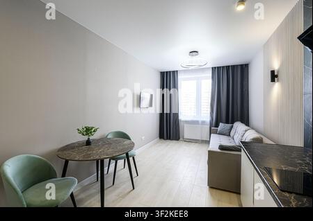 appartement moderne doté d'un coin repas avec table ronde et deux chaises, d'un canapé confortable près de la fenêtre et d'un coin cuisine épuré Banque D'Images