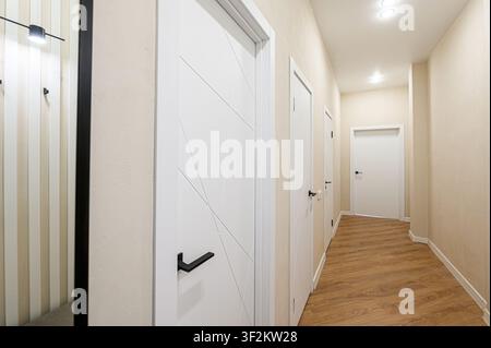 Vue intérieure d'un couloir comportant plusieurs portes blanches avec poignées noires. Des murs neutres et un revêtement de sol aspect bois apportent une esthétique propre et moderne Banque D'Images