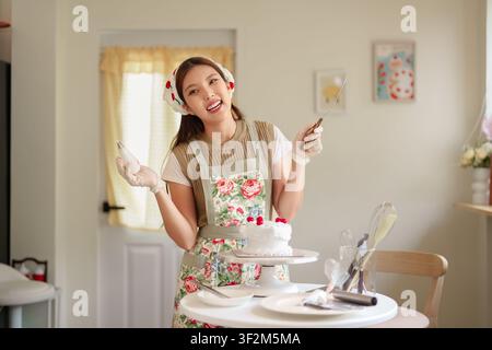 Décoratrice de gâteaux joyeuse présentant un dessert fait maison avec des outils de décoration dans un espace de travail de cuisine baigné de soleil Banque D'Images