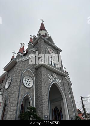 6 avril 2025, Hué, Vietnam – Vue dramatique en contre-plongée de la façade en pierre néo-gothique d'une église catholique avec des croix, des rosaces et des détails ornés sous un ciel nuageux. Banque D'Images