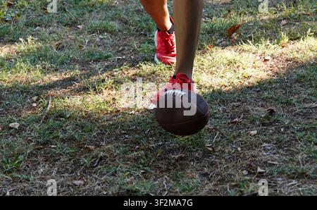 Un pied chaussé de baskets rouges entre en contact avec un ballon de football marron sur l'herbe, le soleil créant des ombres et mettant en valeur la texture du sol Banque D'Images