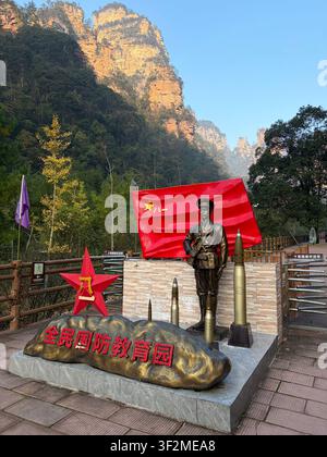 5 novembre 2025, Wulingyuan, Zhangjiajie, Chine – Statue en bronze d’un soldat avec drapeau rouge du PCC, obus d’artillerie et inscription patriotique chinoise devant des piliers karstiques en grès. Banque D'Images