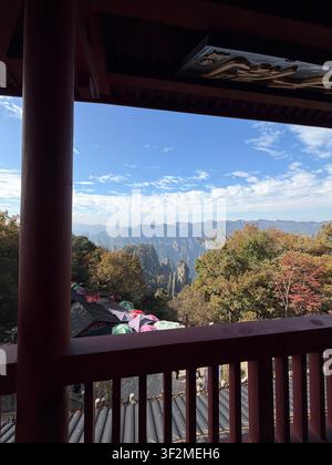 5 novembre 2025, Wulingyuan, Zhangjiajie, Chine – Piliers karstiques en grès de Wulingyuan vus à travers le cadre d’un pavillon en bois avec feuillage d’automne et parapluies colorés. Banque D'Images