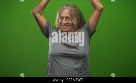 Femme avec les bras levés et souriant dans un fond vert studio, tshirt gris visible et pose expressive montrant les bras levés et le rire ; Joy celeati Banque D'Images