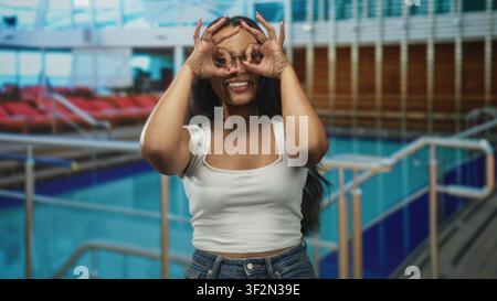 Femme brune dans un bureau blanc et un Jean, mains formant des jumelles sur les yeux tout en souriant à une terrasse de la piscine intérieure avec chaises longues et balustrades ; pl Banque D'Images