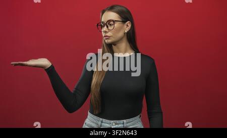Femme avec lunettes tenant la paume vers le haut et levant l'index dans une pose étudiée en rouge, taille en jean visible ; instruction affirmée. Banque D'Images