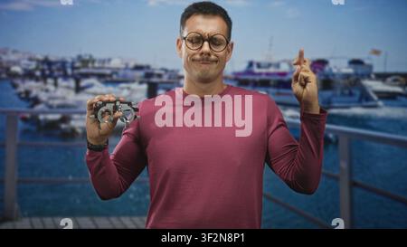 Jeune homme hispanique portant une moustache et des lunettes rondes, tenant un cadre d'essai et croisant les doigts en studio ; chance espérée. Banque D'Images