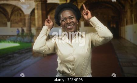 Femme pointe les index vers le haut montrant les mains et le sourire dans un couloir de bâtiment historique ; célébration de joie. Banque D'Images