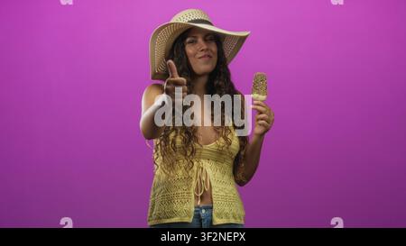 Femme hispanique portant un chapeau de soleil et un haut crochet jaune sourit, tient un popsicle et donne les pouces en studio ; joie d'été. Banque D'Images