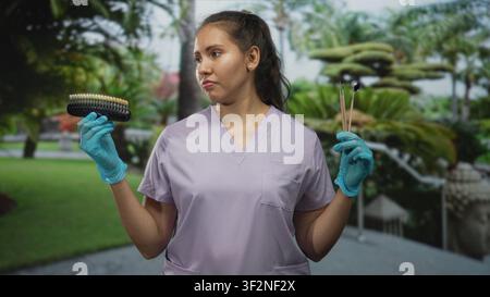 Femme dentiste dans les gommages tient le guide de teinte dentaire et les miroirs buccaux dans les mains gantées dans la forêt ; évaluation des préoccupations. Banque D'Images