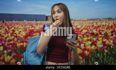 Jeune femme main à bouche mangeant une collation tout en tenant un smartphone et un sac à dos dans un champ de tulipes coloré ; contentement de la jeunesse. Banque D'Images