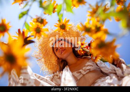 Femme cueillant des fleurs de rudbeckia à l'aide d'une sécateur dans un jardin en été. Portrait vu d'en bas. Jardinière élégante portant un chapeau de paille devant le ciel Banque D'Images