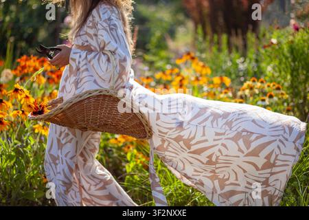 Femme se promenant dans un jardin d'été en cueillant des fleurs de rudbeckia avec un panier de récolte. Jardinière portant une robe de chambre longue et élégante Banque D'Images