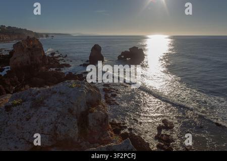 La lumière du soleil se reflète sur l'eau au rivage rocheux. Les vagues s'écrasent contre les rochers tandis que la côte s'étend au loin. C'est un matin lumineux Banque D'Images