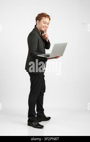 Prise de vue en pleine longueur d'un homme d'affaires asiatique joyeux et enthousiaste portant un costume et tenant un ordinateur portable sur un fond isolé Banque D'Images