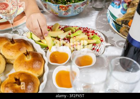 Les gens se rassemblent pour célébrer Rosh Hashanah. Une table est dressée avec des pommes, du miel et du pain challah rond. Des mains tendues attrapent les aliments sucrés symbolisant les vœux pour une bonne année. Banque D'Images