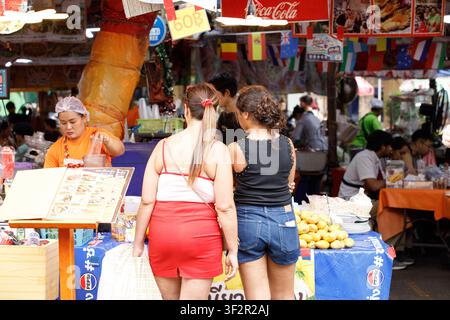 BANGKOK, THAÏLANDE - 12 JUILLET 2026 : Touristes féminines achetant du riz gluant au mango sur un marché en plein air à Bangkok, vue arrière. Banque D'Images