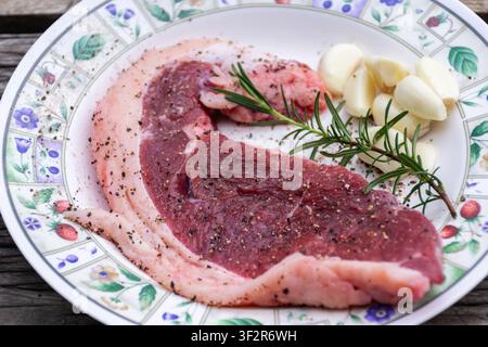 Steak de porc cru frais assaisonné de poivre noir, d'ail et de romarin sur une assiette en céramique, prêt pour la cuisson, le barbecue ou la préparation d'un repas maison Banque D'Images