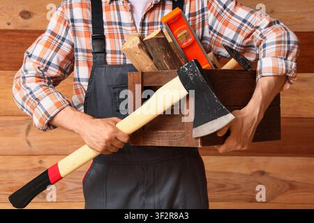 Homme en chemise à carreaux et salopette tenant une caisse en bois remplie d'une hache, d'un niveau et de bois de chauffage contre un fond en panneaux de bois Banque D'Images