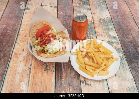 Sandwich au doner chargé avec de la laitue et des sauces, accompagné de frites ondulées et d'une boisson en canette sur des planches en bois. Banque D'Images
