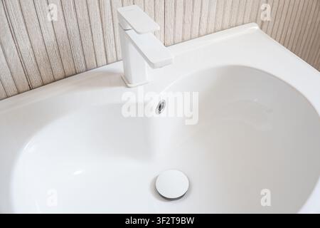 Prise de vue en contre-plongée d'un lavabo en porcelaine blanc immaculé doté d'un robinet élégant, d'un bouchon d'évacuation central et d'un trou de trop-plein contre un fond texturé clair Banque D'Images