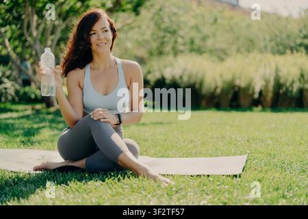 Femme brune souriante en tenue de sport tenant une bouteille d'eau assise sur un tapis de yoga après un entraînement en plein air dans le parc Banque D'Images