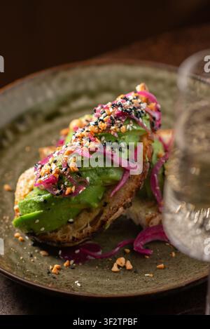 Près-plan d'un toast gourmand à l'avocat garni d'oignons rouges marinés et de graines de sésame, avec un verre de vin pétillant au premier plan. Banque D'Images