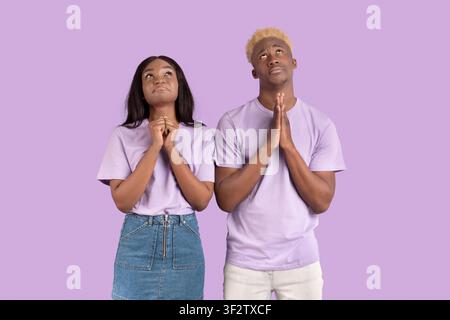 Couple religieux noir mettant les mains ensemble dans la prière, levant les yeux vers le haut, demandant à Dieu de l'aide sur fond lilas Banque D'Images