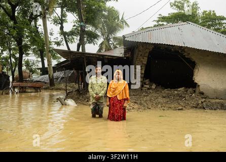 Banshkhali Upazila, Division de Chittagong, Bangladesh - 12 juillet 2026 : Vue d'un couple debout dans les eaux d'inondation devant leur maison endommagée après de fortes Banque D'Images
