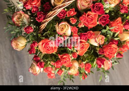 Vue de dessus en gros plan de roses colorées disposées dans un panier en osier avec la anse visible. Une lumière chaude et un fond brun clair créent une fleur naturelle Banque D'Images