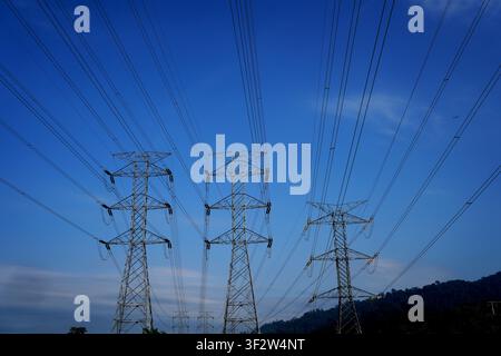Trois poteaux de lignes à haute tension et câbles électriques aériens sur un ciel bleu profond et dégagé avec des collines au loin Banque D'Images
