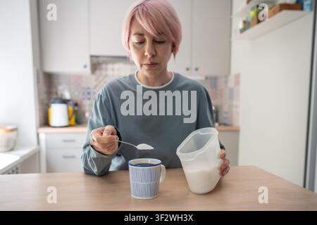 Femme mûre ajoutant une cuillerée de sucre à un café matinal aromatique, consommation incontrôlée d'édulcorants. Banque D'Images
