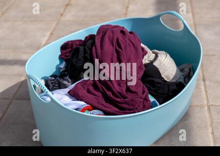 Panier à linge en plastique bleu rempli de vêtements sales variés posé sur un sol carrelé sous la lumière naturelle du jour. L'image illustre les tâches ménagères quotidiennes Banque D'Images