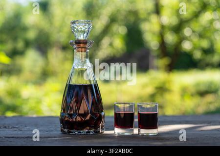 Liqueur de cerise maison dans un décanter et deux verres à liqueur. Carafe en cristal remplie de liqueur de cerise rouge foncé à côté de deux verres à liqueur remplis sur un bois rustique Banque D'Images