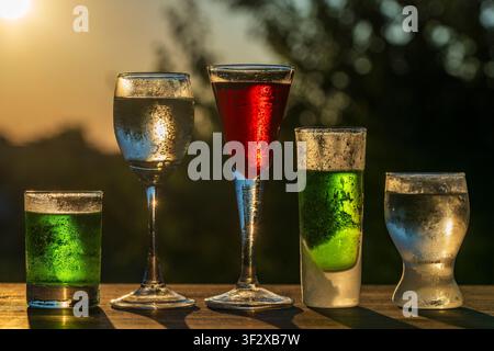 Ensemble de diverses boissons alcoolisées, liqueurs et shots dans des verres dépareillés sur une table en bois en plein air, gros plan. Boissons vert vif, rouges et claires Banque D'Images