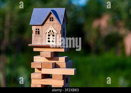 Maison en bois jouet au sommet d'une tour instable faite de blocs de Jenga en plein air, gros plan. Métaphore du risque immobilier, de l'instabilité du marché immobilier, du prêt immobilier Banque D'Images