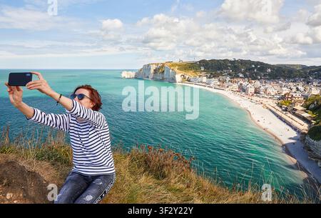Femme tenant un smartphone pour prendre un selfie au sommet d'une falaise avec vue panoramique sur la plage et la ville d'Etretat en Normandie. Banque D'Images