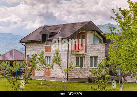 21 mai 2026, Mojkovac, Monténégro : Maison moderne arborant le drapeau national sur le balcon, entourée de nature verdoyante et de montagnes lointaines Banque D'Images