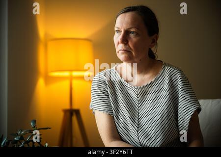 Femme d'âge moyen plongée dans ses pensées en étant assise dans un salon confortable. Thème de réflexion, résolution de problèmes, préoccupation et bien-être émotionnel. Mise au point sélective Banque D'Images