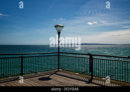 Une lanterne avec des rampes au bout d'un ponton sur une mer bleu-vert avec des reflets de soleil et une pointe de terre au loin, Boscombe Pier, Bournemouth. Banque D'Images