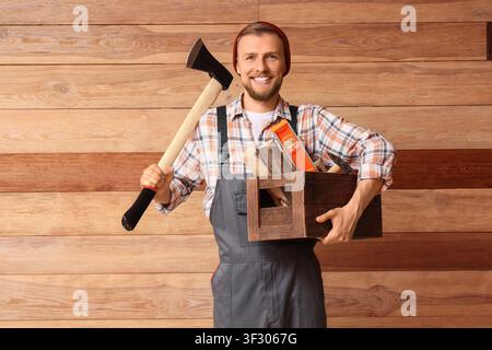 Jeune charpentier souriant en salopette tenant une hache et une boîte à outils en bois avec des outils devant un fond de panneaux de bois, l'homme se tient confiant contre Banque D'Images