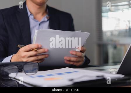 Gros plan d'un homme d'affaires tenant et lisant un rapport d'affaires. Lunettes sur le bureau au premier plan. Cadre examinant des documents financiers, juridique co Banque D'Images
