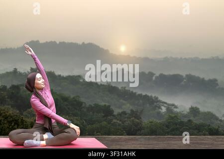 Une jeune femme musulmane indonésienne d'Asie du Sud-Est portant un hijab pratique le yoga et des étirements sur un tapis en plein air devant un magnifique paysage montagneux brumeux Banque D'Images