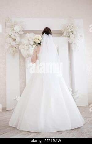 Mariee en robe de mariée blanche et voile tenant un bouquet devant une arche fleurie Banque D'Images