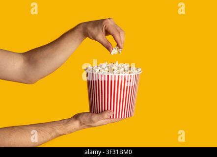Gros plan sur les mains mâles tenant le godet jetable avec du pop-corn sur fond orange, espace de copie. Panorama Banque D'Images