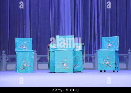 Sur une scène d'opéra Pingju, des chaises et des tables recouvertes de soie turquoise brodée de fleurs de prunier blanches sont soigneusement disposées contre un cuif bleu profond Banque D'Images
